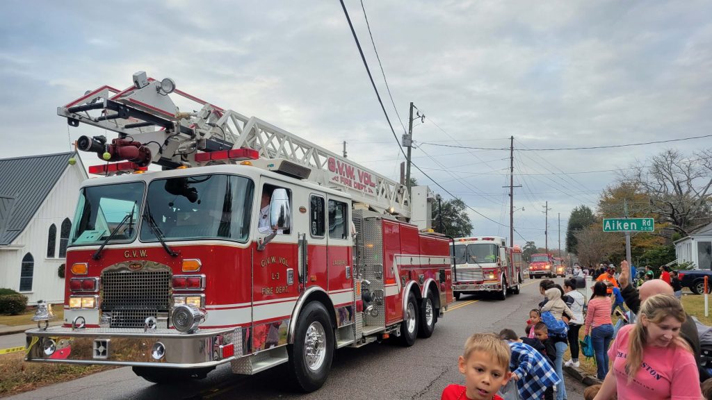 A large red fire truck drives along a street lined with people watching a parade. Children and adults sit on the curb, and more fire trucks are visible in the background. The sky is overcast, and a church is visible on the left side.