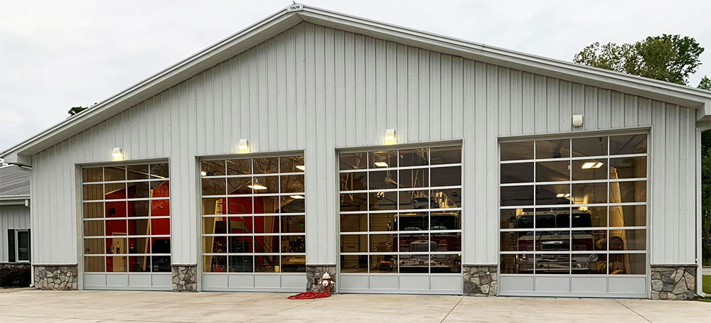 A large fire station with four glass garage doors, each housing a fire truck. The building is light gray with stone accents at the base and is situated on a wide concrete driveway. Trees are visible in the background.