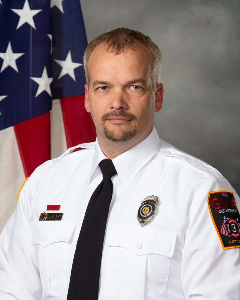 A man in a white uniform with patches on the sleeves stands in front of a U.S. flag. He has short hair and a mustache, wearing a black tie, looking directly at the camera. The background is a neutral gray.