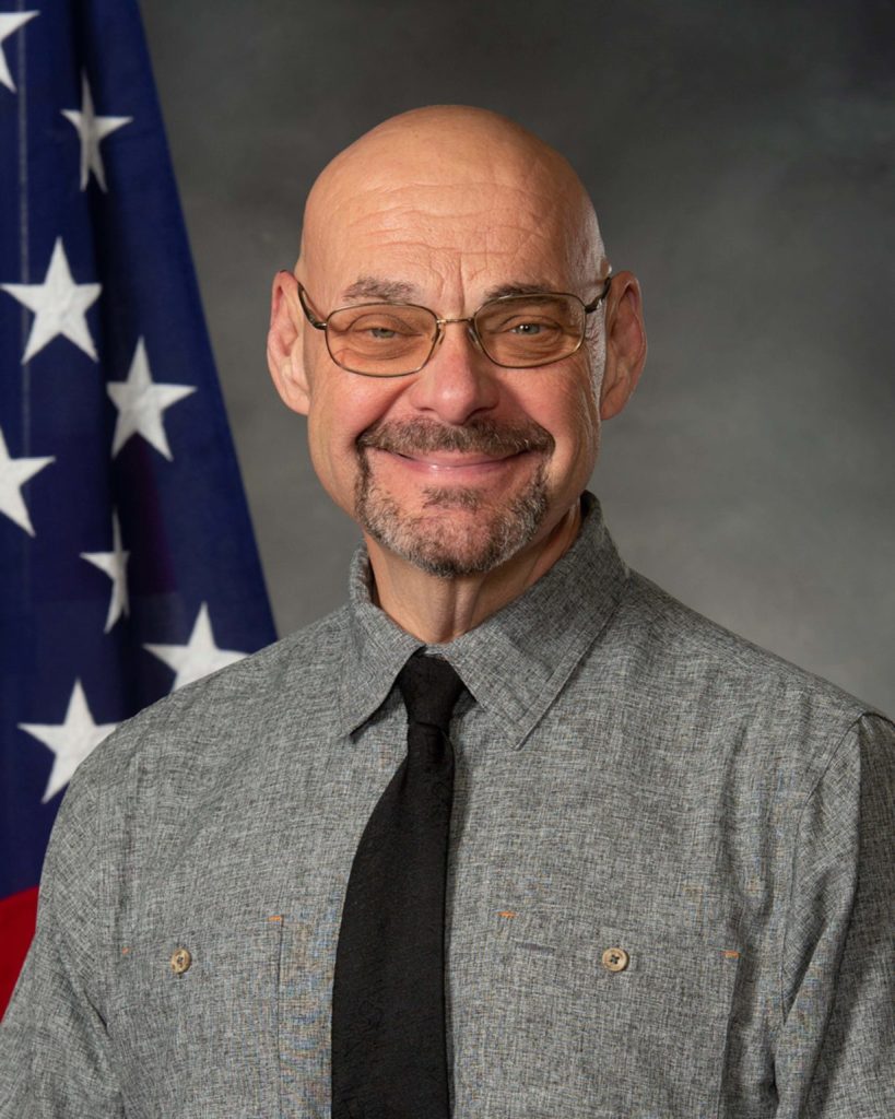 A person wearing glasses and a gray button-up shirt with a black tie is smiling. They stand in front of an American flag and a gray background.