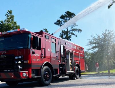 A red fire truck from the GVW Fire Dept is stationed on a sunny road, spraying water into the air. Tall trees and a few people stand in the background, observing the scene.
