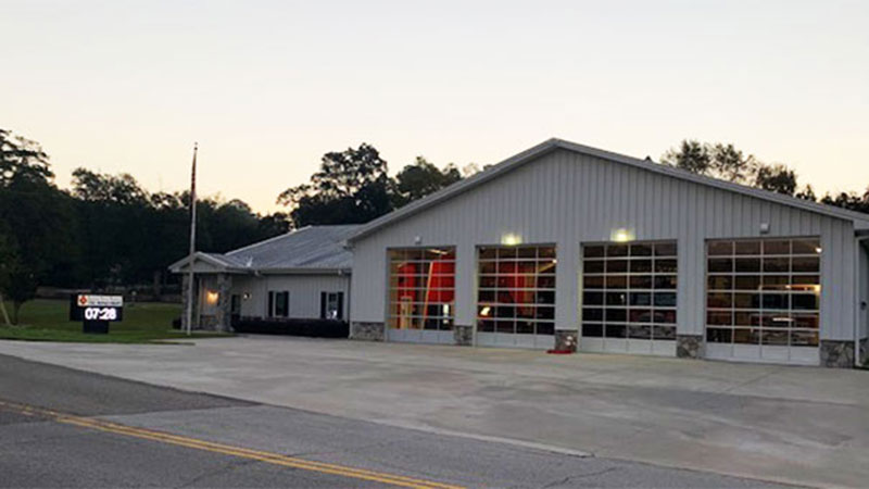 A fire station with a corrugated metal exterior and multiple garage doors, surrounded by trees under a clear sky. A digital sign displays "07:28" near the entrance. The area is quiet and the road in front is empty.