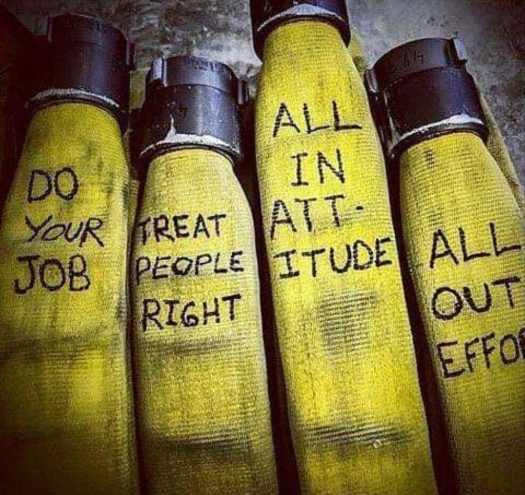 Four yellow fire hoses are lined up side by side, each with a motivational phrase written on them: "Do Your Job," "Treat People Right," "All in Attitude," and "All Out Effort." The background is dark and muted.