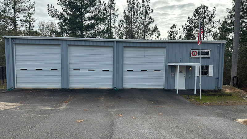 A small fire station with three closed garage doors and a blue-gray exterior, surrounded by trees. An American flag is displayed on a pole in front, and a sign bearing a fire department emblem is visible above the entrance.