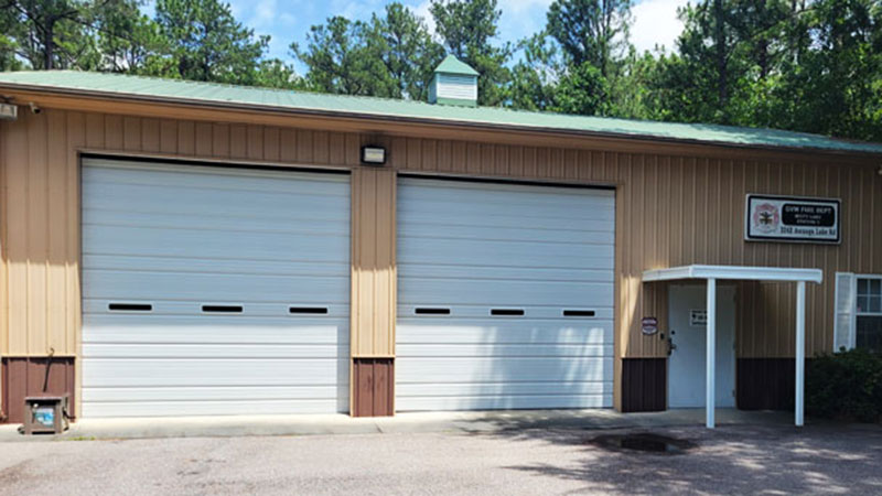 A beige garage building with two large white roll-up doors and a small covered entrance. There is a sign near the roof with text on it. The scene is set against a backdrop of tall green trees under a blue sky.