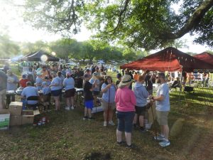 A large outdoor gathering with people standing in line near red and black tents. Many are wearing casual clothing and hats. The event takes place in a grassy area with trees providing shade. Sunlight filters through the leaves, creating a bright atmosphere.