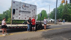 Four people stand on a street next to a trailer with a large banner that says "Bridgestone" and other logos. A girl on the left is holding a flag. Trees and a white vehicle are in the background.