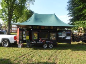 A mobile information trailer with a green canopy is set up in a grassy area. It displays various educational materials and has open panels showing maps and posters. A white pickup truck is parked beside it, and there's a foldable chair nearby.