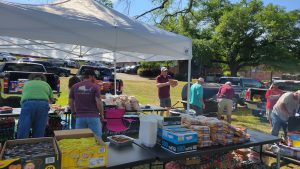 People are gathered under a canopy tent at an outdoor event. Tables are laden with bags of chips, loaves of bread, and other food items. Several individuals are preparing or organizing the items. Cars are parked in the background under sunny skies.
