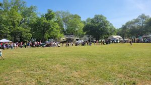 A large group of people is gathered on a grassy field under a clear blue sky. Trees and small white buildings line the background. Tents are set up at the far end, and people are spread out across the open space.