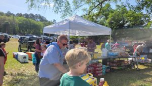 People gathered outdoors under a white canopy tent, surrounded by coolers, drinks, and snacks. A sunny day with trees and cars in the background. Some individuals are interacting, while others are preparing or serving food and drinks.
