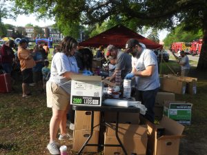 People are gathered outdoors at a community event, standing by tables and handling canned goods. Cardboard boxes and supplies are visible. A red tent and fire trucks are in the background, with trees and buildings nearby.