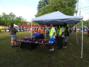 A group of people stands under a blue canopy tent at an outdoor event. The table beneath the tent displays various items. Other tents and a crowd are visible in the background on a grassy area. Trees surround the scene.