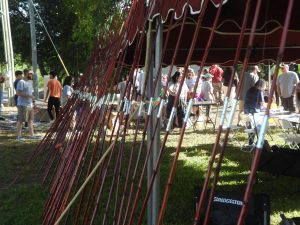 A row of fishing rods is lined up under a canopy at an outdoor event. People are gathered in the background, engaging in various activities under the shade of trees. The ground is grassy, and sunlight filters through the canopy.