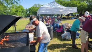 A man grills hot dogs on a large barbecue in a park. People are lined up under a white tent in the background, with tables displaying buns and supplies. Trees and cars are visible in the distance. It appears to be a sunny day.