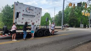 A group of people stands around and on a trailer with a large sign displaying the Bridgestone logo and the phrase "Solutions for your journey." The trailer is parked on a street near a railroad crossing with several trees lining the background.
