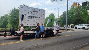 People stand in front of a trailer featuring a Bridgestone banner, with logos and the phrase "Solutions for your journey." They appear to be participating in an event on a road. There are trees and a railroad crossing in the background.