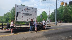 People stand in front of a trailer with a "Bridgestone" banner at an outdoor event. The setup is near a road, surrounded by trees and a railroad crossing. Some participants appear to be holding items, and a few people have microphones.