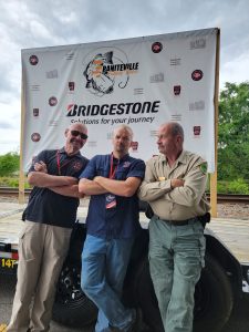 Three men stand with arms crossed in front of a banner on a platform. They wear casual clothing and identification badges. The banner features logos including Bridgestone. A railway track and greenery are visible in the background.