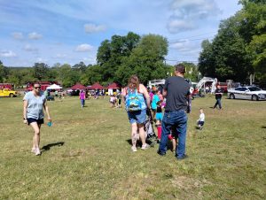 People are gathered outdoors at a park event. Tents and emergency vehicles are in the background. Families and children are scattered across the grassy area, enjoying a sunny day with scattered clouds. Some people are walking, and others are sitting.