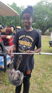 A young person stands outdoors holding a small net containing fish. They wear a "North Augusta Football Jackets" t-shirt. A golf cart and trees are visible in the background.