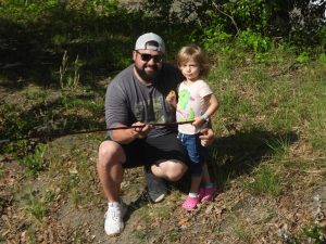 A man wearing a cap and sunglasses crouches on a grassy area, holding a fishing rod with a little girl standing beside him. The girl wears a T-shirt with a colorful dinosaur and pink shoes, and they both smile at the camera.