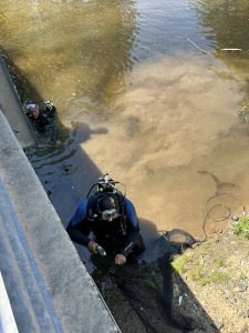 Two scuba divers in murky water near a bank; one is holding a device. They are partially shaded, with a fence on the left and vegetation on the right. The water shows some debris and shadows.