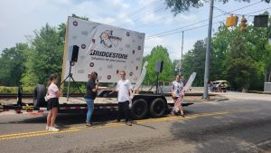 People are gathered around a trailer with a large banner that reads "BRIDGESTONE Solutions for your journey." The setup includes microphones and speakers. Trees and traffic lights are visible in the background.
