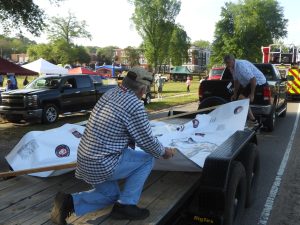 Two men set up a float on a trailer, adjusting a banner with various logos. A fire truck and pickup trucks are in the background, along with colorful tents and trees in a park-like setting.