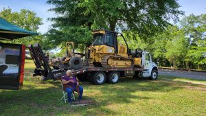 A woman sits on a foldable chair next to a flatbed truck carrying a large yellow bulldozer. The truck is parked on grassy ground under trees, with blue skies visible.