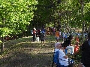 People gathered in a park with fishing rods amid trees. A person sits in a blue chair in the foreground, while others stand or walk near a small body of water. It's a sunny day, and the setting is relaxed and green.