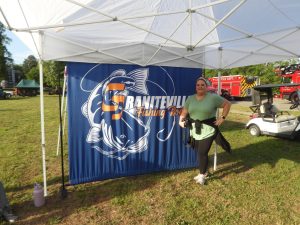 A person stands smiling next to a large blue banner under a canopy. The banner features a fish graphic and text that reads "Graniteville Fishing Rodeo." There are emergency vehicles in the background on grass.