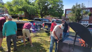 People grill hot dogs on several large grills in a grassy area with trees and a brick building in the background. Folding chairs and pickup trucks are nearby. It's a sunny day, and the group is casually dressed.