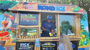 A person serves shaved ice from a tropical-themed food truck with vibrant decorations. The truck has a sign reading "We are nutritious & delicious." A customer stands in front, waiting. Bright, sunny day with trees in the background.