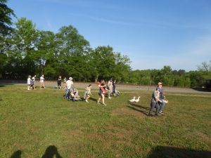 A group of people, including children and adults, walk across a grassy field under a clear blue sky. Some push strollers. Two ducks are nearby. Trees line the background, and a dirt path is visible.