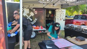 People are preparing food at a barbecue stall. A man in an apron and another in a cap are working at a table with a logo in the background. A woman with long hair is writing on a whiteboard menu. Plates of food and a red truck are nearby.
