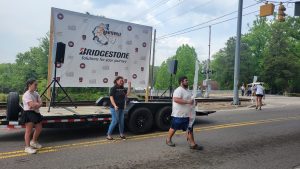 A group of people walk along a road beside a trailer with a Bridgestone banner. One person speaks into a microphone, while others carry signs. Trees and a railroad crossing are visible in the background.