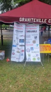 A red canopy with "Graniteville" printed on it shelters two sponsor banners. The banners list various logos, including Bridgestone and Reliable. A yellow sign partially reads “EGISTRATION.” A cardboard box sits on the grass.