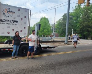 Two people holding signs stand near a trailer with a Bridgestone logo at a street intersection. A railroad crossing is visible in the background. Trees line the street, and a person wearing a hat walks away in the distance.
