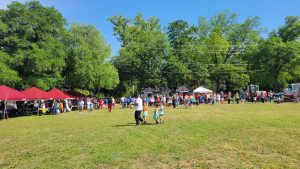A community event in a park with many people walking and gathering. Red and black tents are set up in the background. Trees surround the open grassy area under a clear blue sky.