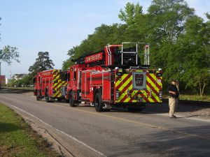 Two red fire trucks with high visibility chevron patterns parked on a road near railroad tracks. A person stands nearby, holding a radio. Trees and houses are visible in the background under a clear sky.
