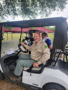 Two people sit in a golf cart under a tree. The person in front, wearing a beige shirt with a patch, smiles at the camera while holding the steering wheel. The other person, wearing a dark shirt, adjusts something in the back. Tents are visible in the background.