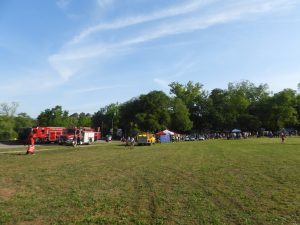 A grassy field with a few people walking. Two fire trucks are parked on the left. There are several tents and a crowd gathered under the trees in the background. The sky is clear with thin clouds.