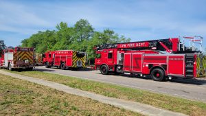 Fire trucks from GVW Fire Dept. and an Engine 2 vehicle are parked along a road with trees in the background. The trucks are bright red with various equipment visible. The scene is in daylight under a clear sky.