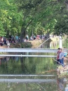 A crowd gathered along a tree-lined riverbank, some sitting in chairs and others standing. A few people fish near the water's edge. The surrounding greenery is reflected in the calm water, creating a serene outdoor scene.