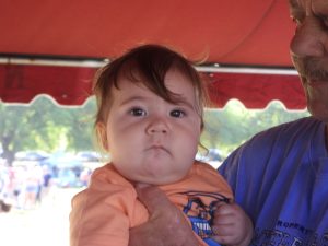 A baby with a tuft of hair on their forehead is being held by an older man under a red canopy. The baby wears an orange shirt with a design. The background is blurred, showing people and greenery.