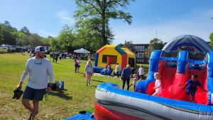 A sunny outdoor event with children playing on colorful inflatable slides and bounce houses. Adults are supervising, and there are tents and more people in the background. A man in a white shirt and cap walks in the foreground, holding his shoes.