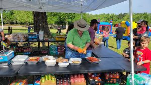 A man in a green shirt and hat prepares food on a table under a canopy at an outdoor event. Various food items and drinks are displayed on tables. Families and children are visible in the background near inflatable attractions.