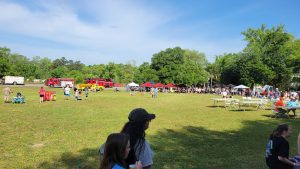 A community event in a grassy field with several people gathered. Two red fire trucks and a yellow vehicle are parked nearby. Tents and tables are set up, with many attendees around. The sky is clear and blue.