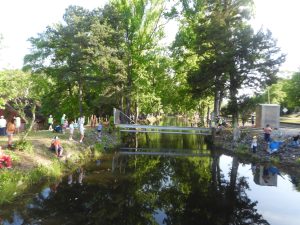 A serene canal surrounded by lush green trees, with people scattered along the banks and a small bridge crossing over the water. Some individuals appear to be fishing, while others are relaxing and enjoying the natural setting.